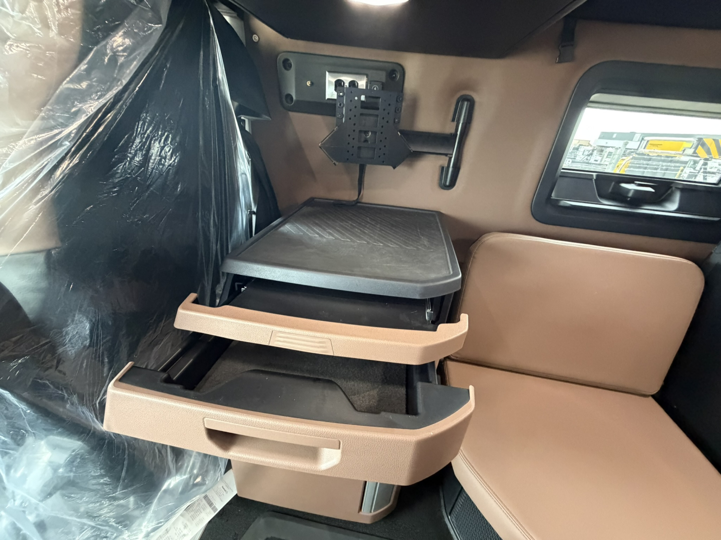 Truck cab interior showing two-folding tan plastic storage trays pulled out, beige leather seat, monitor bracket, and window with stickered view.