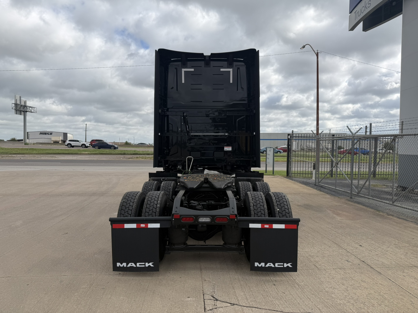 Mack PR64T76H truck rear view showing fifth wheel and dual rear tires with black cab portion, yard fence, and cloudy sky.