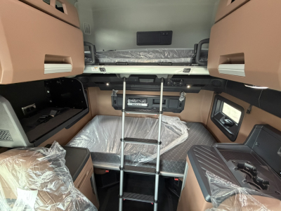 Cab interior of a new Mack PR64T76H truck showing bunk bed area with ladder, upper and lower compartments, beige and black finishes, plastic wrap on seats.
