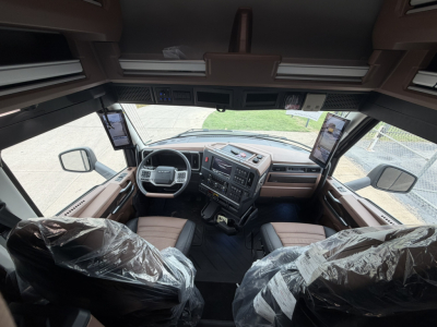 Interior view of a Mack truck cab from cockpit perspective showing steering wheel dashboard and two front seats wrapped in plastic.