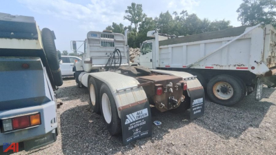 Truck rear view showing fifth wheel and dual rear axles of a Mack E7 tractor chassis in a gravel lot with other trucks in background.