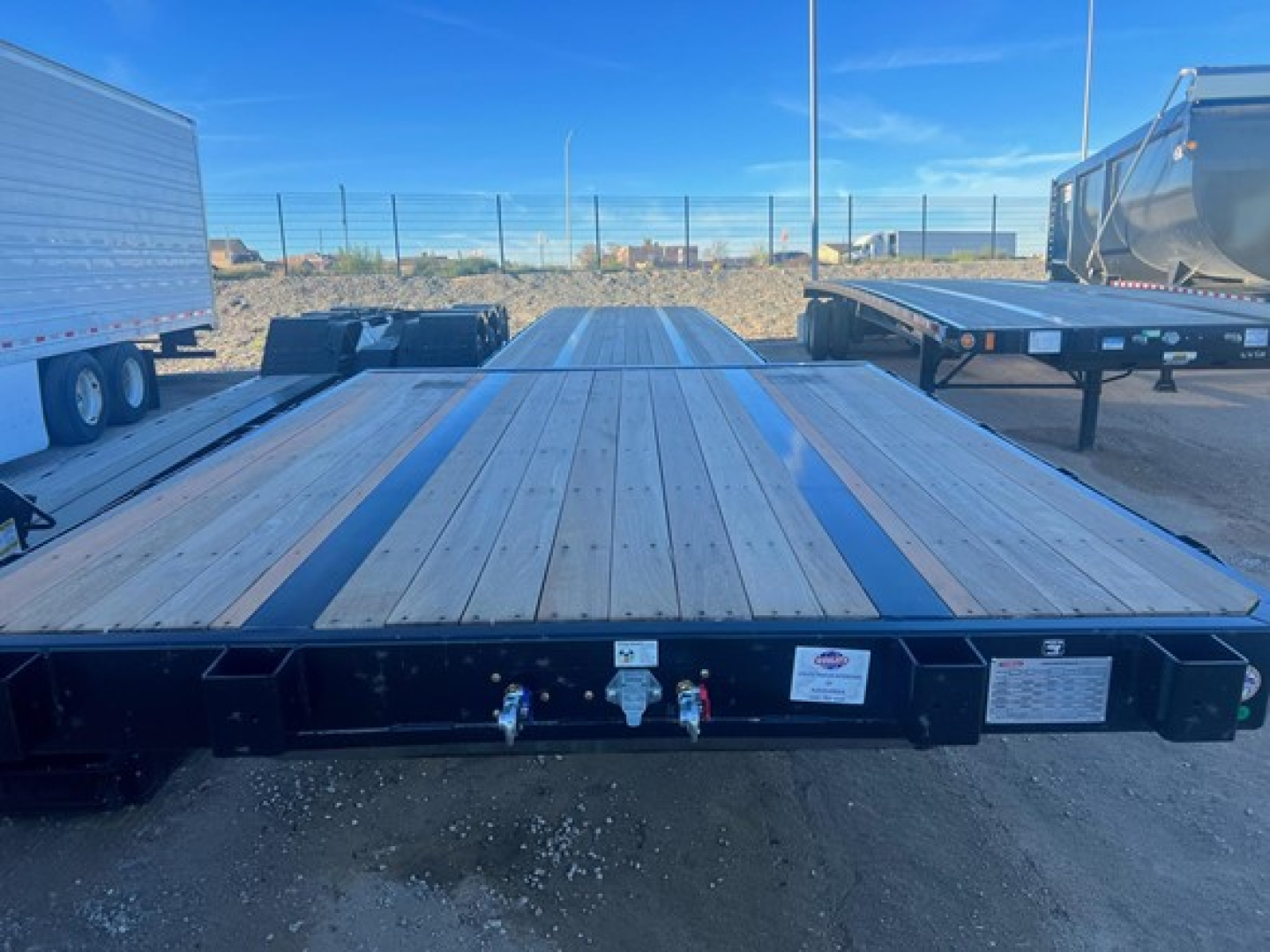 Flatbed trailer with a wooden deck, seen from the front, with blue sky. Visible label plates and coupler area at the front edge.