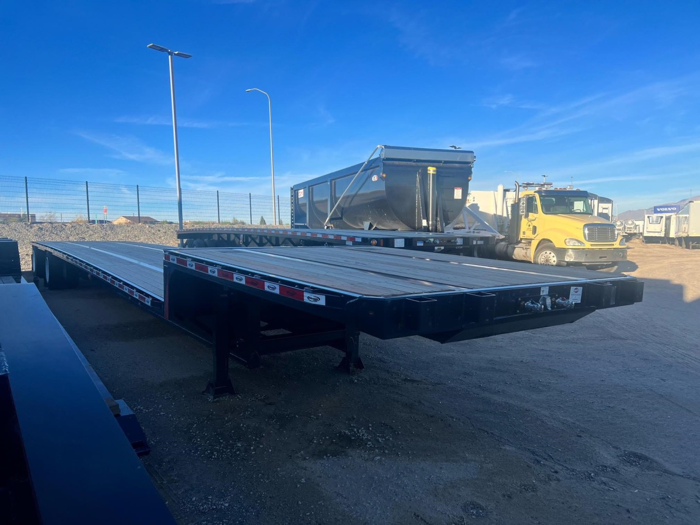 Heavy-duty steel drop deck trailer with long flatbed in a yard, with helper equipment and a dump truck in the background.