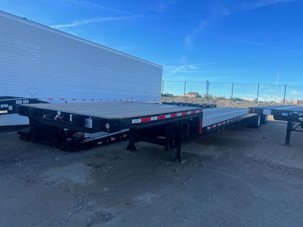 Trailer deck showing low-profile flatbed with black steel frame and drop deck; long tandem axles at rear; reflective safety tape along sides; blue sky; dirt ground.