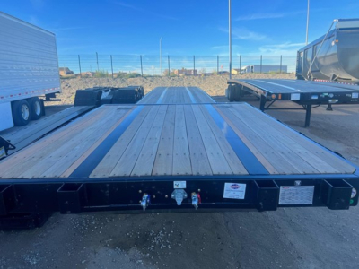 Flatbed trailer with a wooden deck, seen from the front, with blue sky. Visible label plates and coupler area at the front edge.