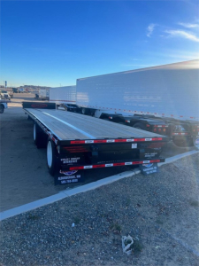 Aluminum drop deck trailer showing 5-axle configuration with steel deck and rear taillights, side reflectors, and stacked trailers in the background.