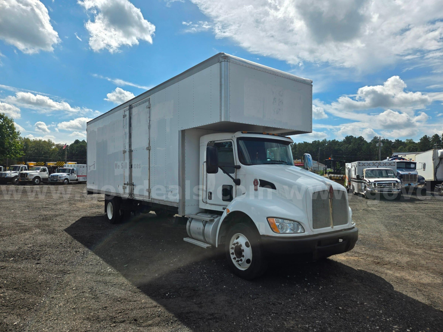 Kenworth box truck in a dealership lot showing a large cargo box mounted over the cab and dual rear wheels.