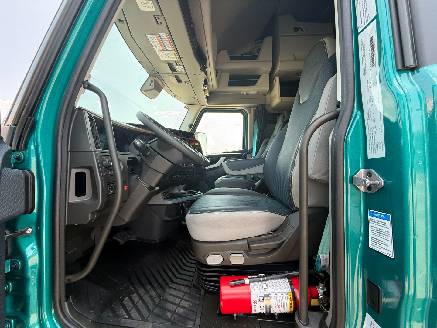 Volvo VNL64T860 cab interior view from open driver door showing steering wheel, dashboard, and driver seat with red fire extinguisher on floor; teal exterior.