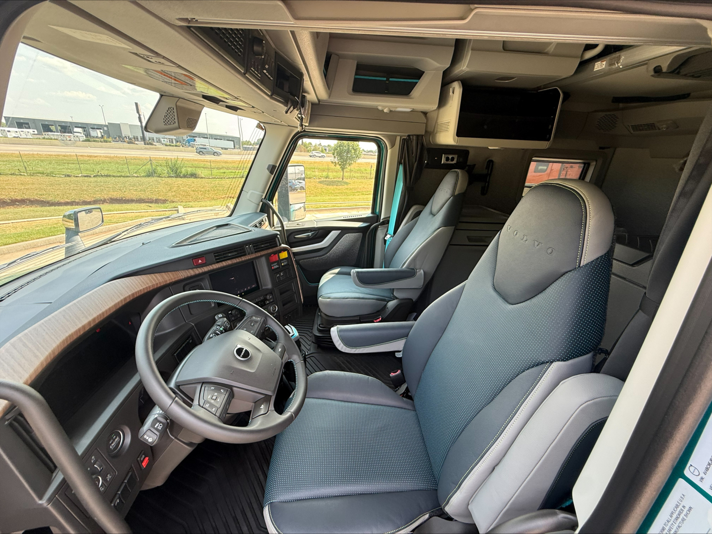 Cab interior of a 2027 Volvo VNL64T860: steering wheel, driver seat and passenger seat, dashboard with digital displays, overhead console, and bunk area visible.