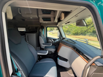 Cab interior of a new Volvo VNL64T860 with gray seats, modern dashboard, steering wheel, and overhead storage. Passenger seat swiveled; view through open door.