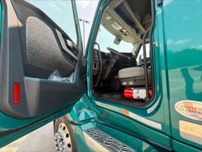 Cab interior of a new Volvo VNL truck with open door, teal exterior, showing driver's seat, steering wheel, and control panel.
