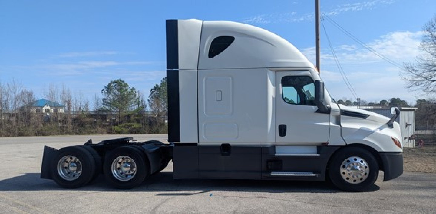 Truck side profile showing 2023 Freightliner Cascadia 126 sleeper tractor with white cab and black chassis, tandem rear axles, daytime parking lot.