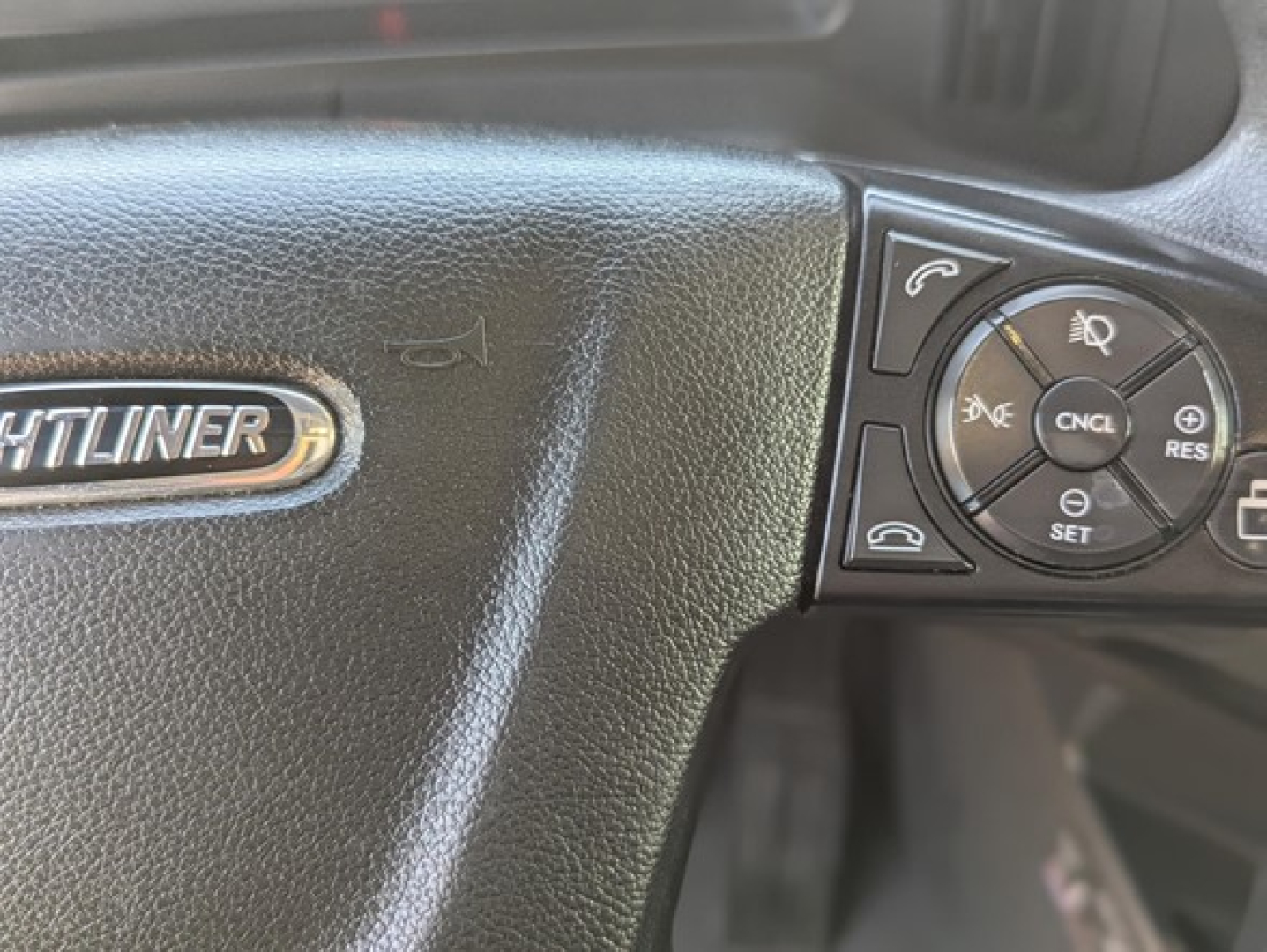 Steering wheel of a Freightliner Cascadia showing part of the dash with control buttons and a 'FREIGHTLINER' badge.
