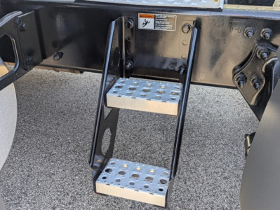 Step ladder under Freightliner Cascadia chassis showing perforated metal steps and black frame, warning label on the frame.
