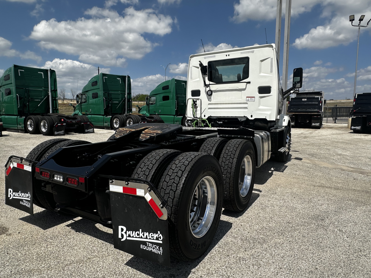 Truck rear view showing seventh axle and mud flaps with Bruckner's Truck Equipment logo; white cab and green trucks in background.