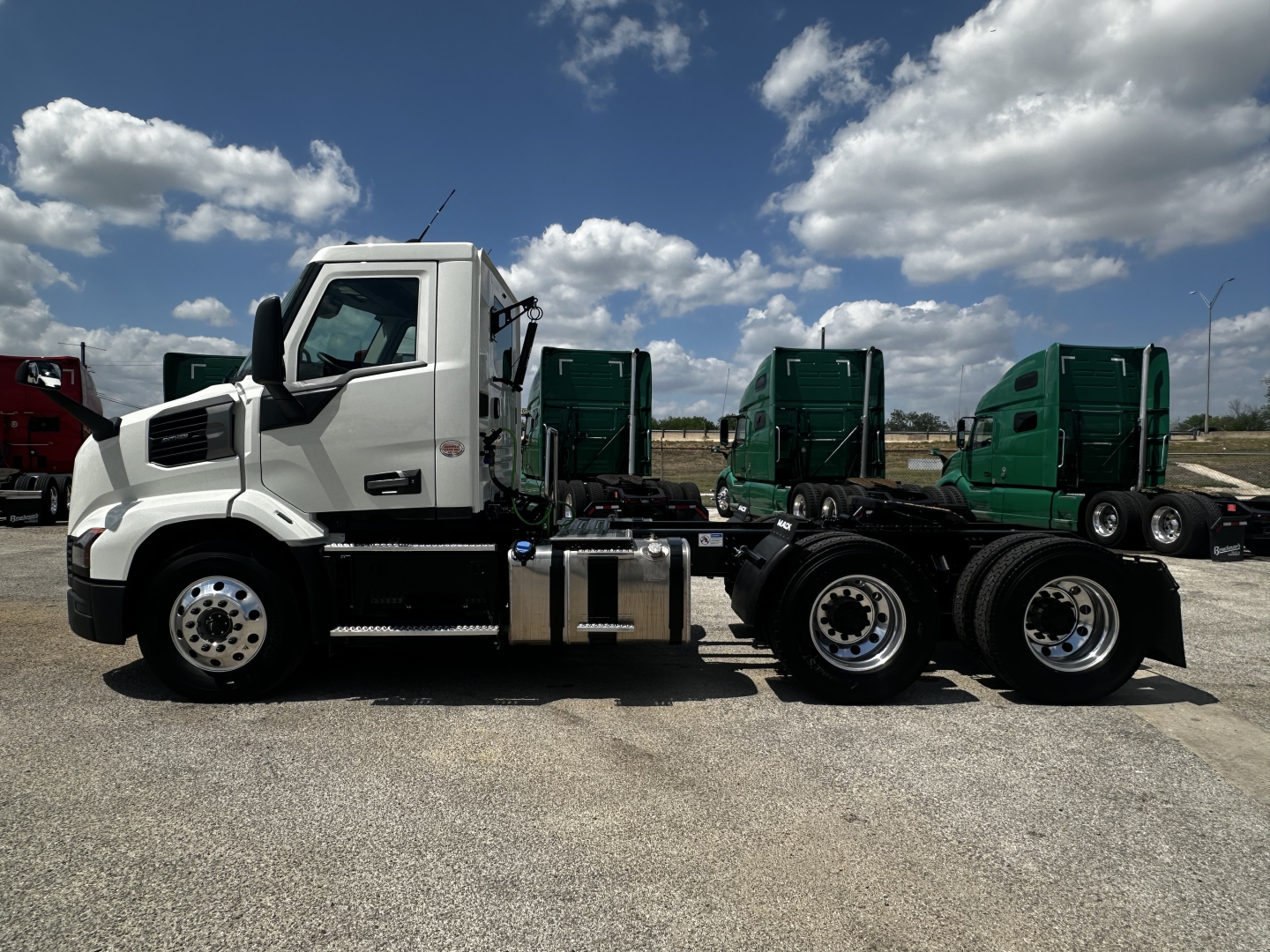 Mack AN64T tractor in white cab profile showing side view with dual rear axles and fuel tanks, chrome exhaust, and green trucks in background.