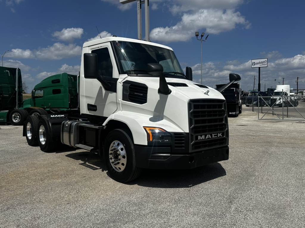 2027 Mack AN64T conventional daycab truck in a dealership lot, white cab, black grille, dual rear axles, with other trucks in background.