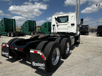 Truck rear view showing seventh axle and mud flaps with Bruckner's Truck Equipment logo; white cab and green trucks in background.