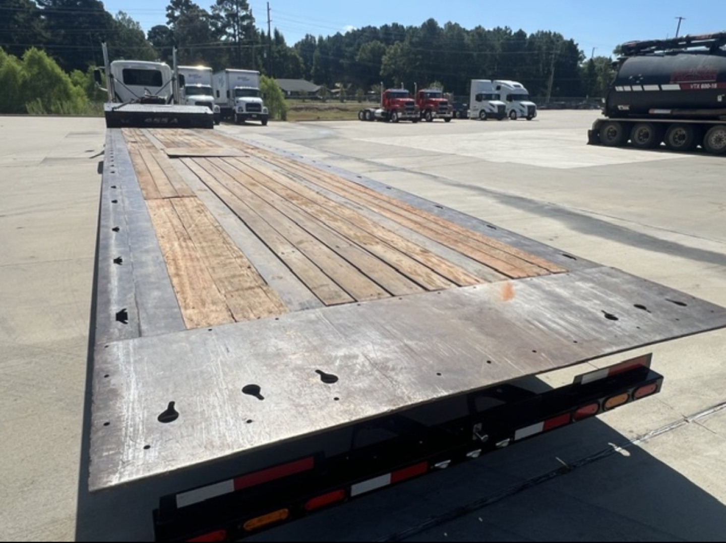 Flatbed trailer bed showing worn wooden deck with metal edges and light reflection; multiple trucks in background.