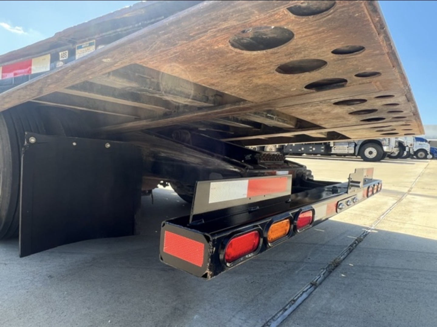 Trailer underside showing landing gear and hydraulic components with circular cutouts in the deck. Visible text includes warning labels and reflective lights on the rear edge.