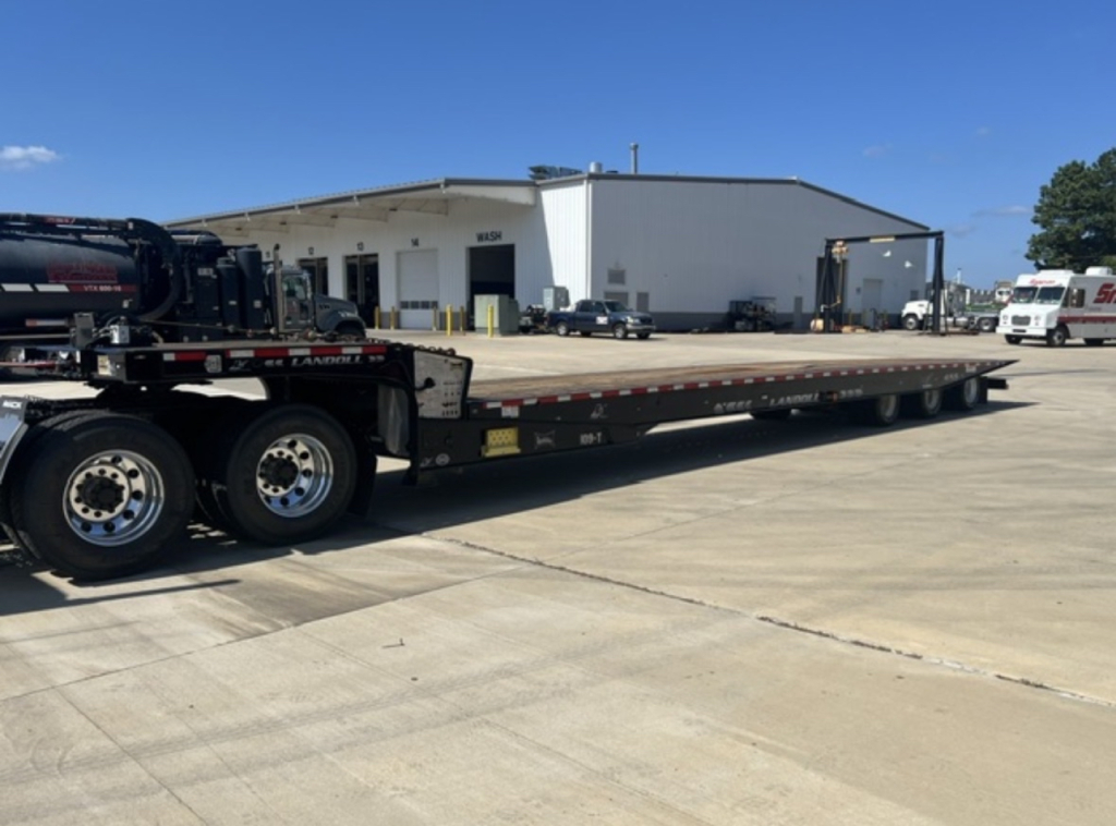 Long flatbed trailer with dual rear axles and hook-lift system. Side shows chain tie-downs and warning labels. Background shows a large warehouse building and other trucks.