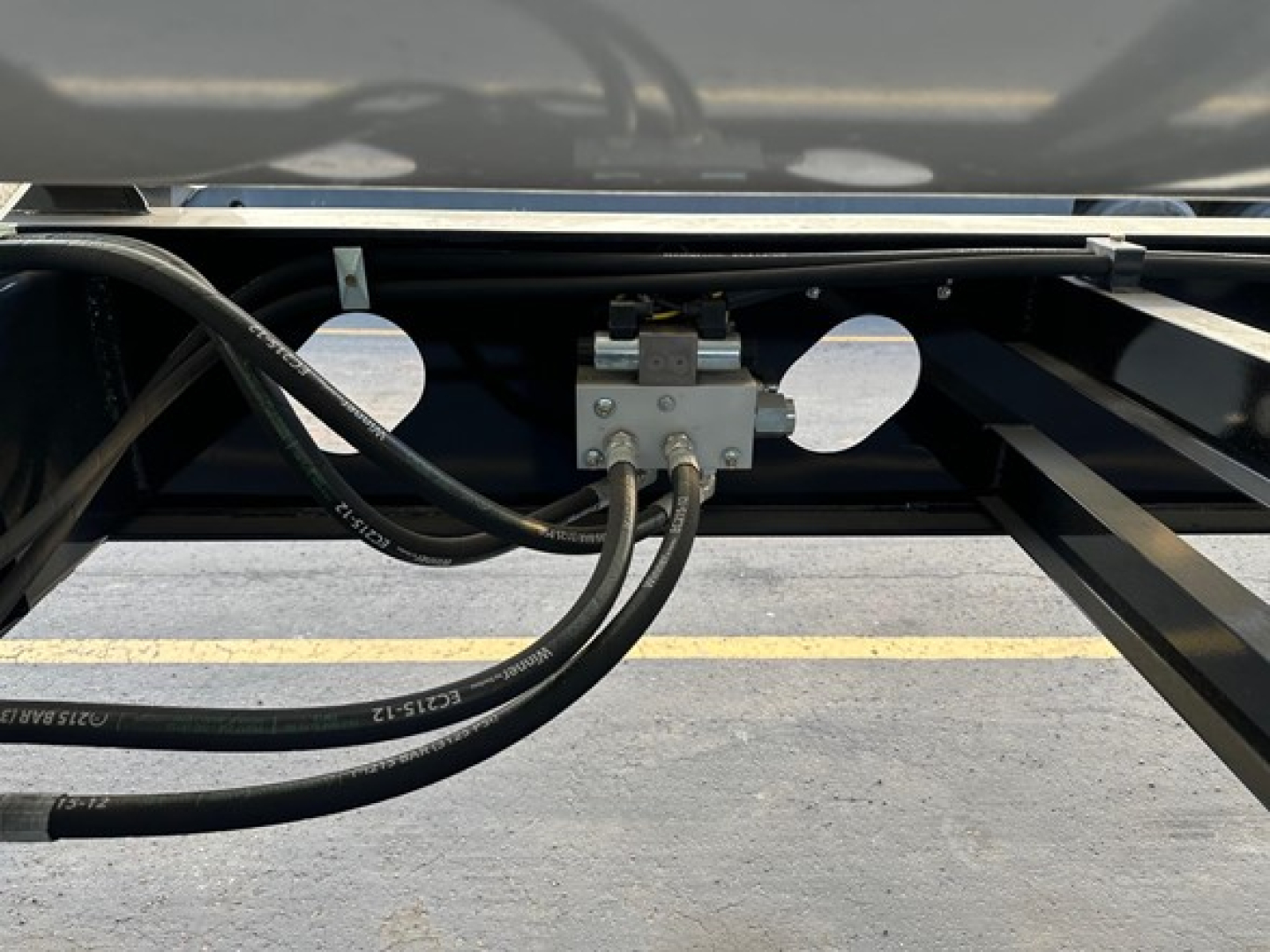 Trailer undercarriage view showing hydraulic lines and a control valve assembly mounted to the frame between circular cutouts.