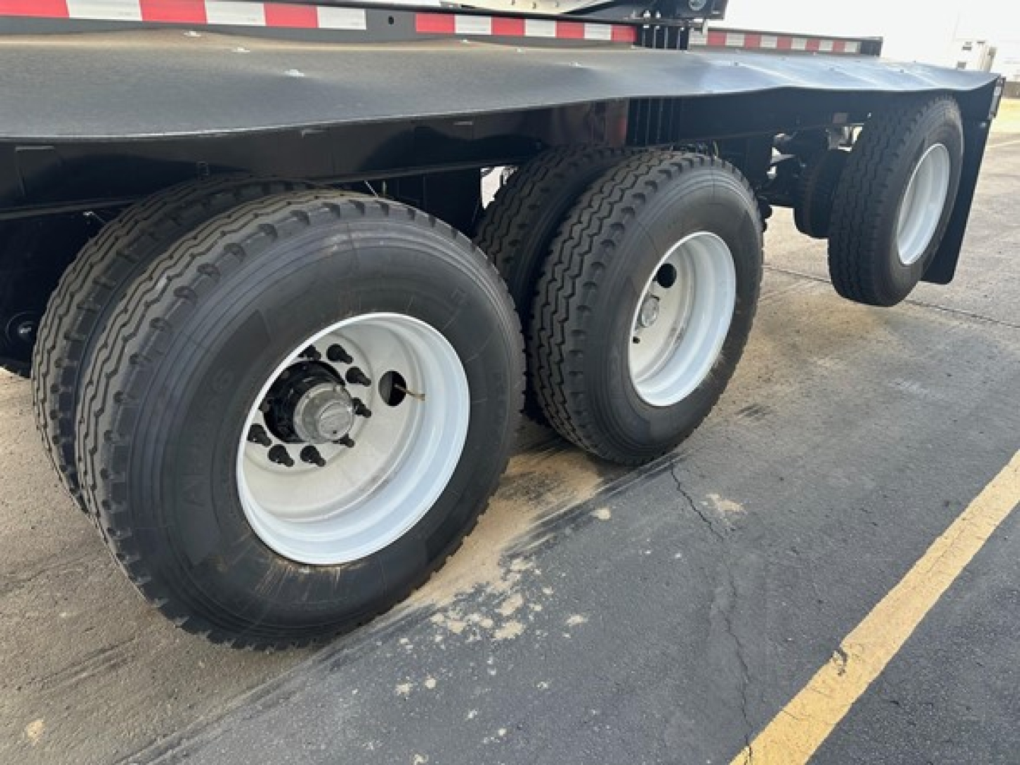 Trailer axle assembly with dual-tire configuration and white rims under a flatbed trailer; visible are three wheels and a portion of the undercarriage.