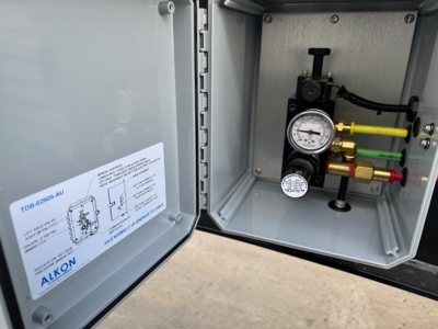 Trailer valve panel inside open access door showing pressure gauge and multiple colored hydraulic lines (green, yellow, red) with fittings and manometer; instruction label on door.