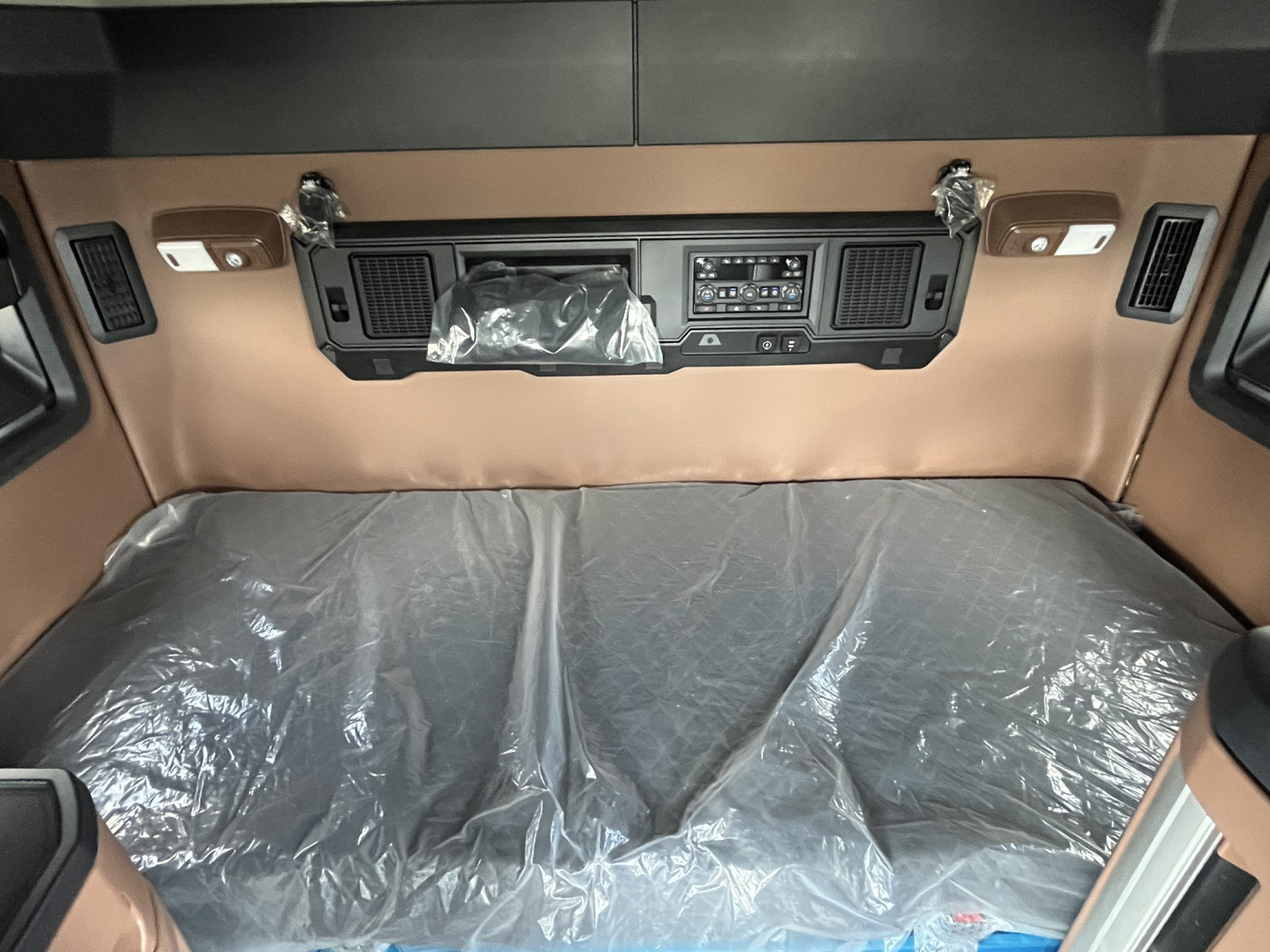 Cab interior of a new Mack AN64T64 truck showing the sleeper cab area with brown dashboard, control panel, and plastic-covered bunk. Visible vents and switches on both sides of the dash.