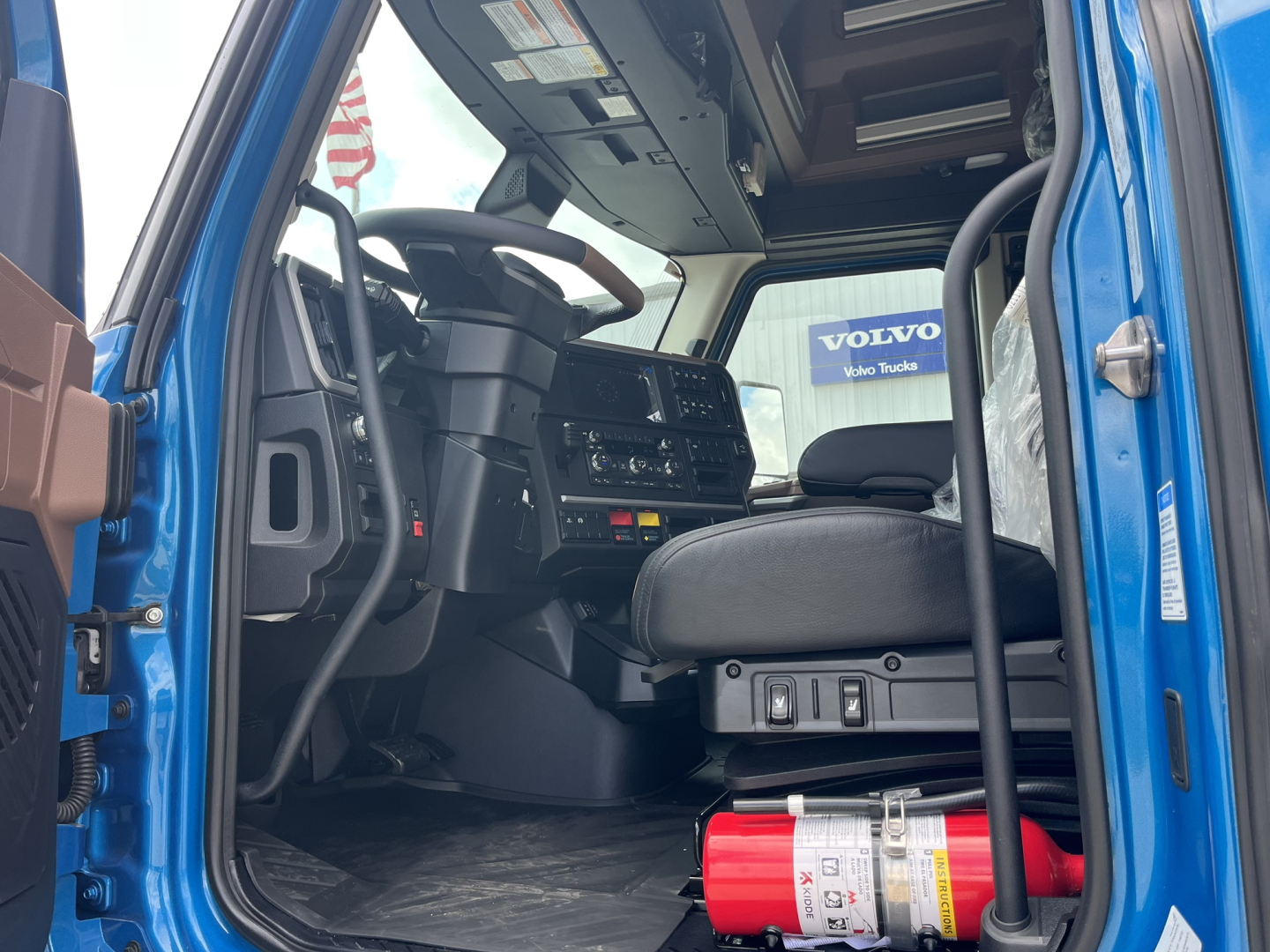 Cab interior of a new Mack AN64T64 truck showing steering wheel, dashboard, and driver seat with a fire extinguisher mounted near the floor.