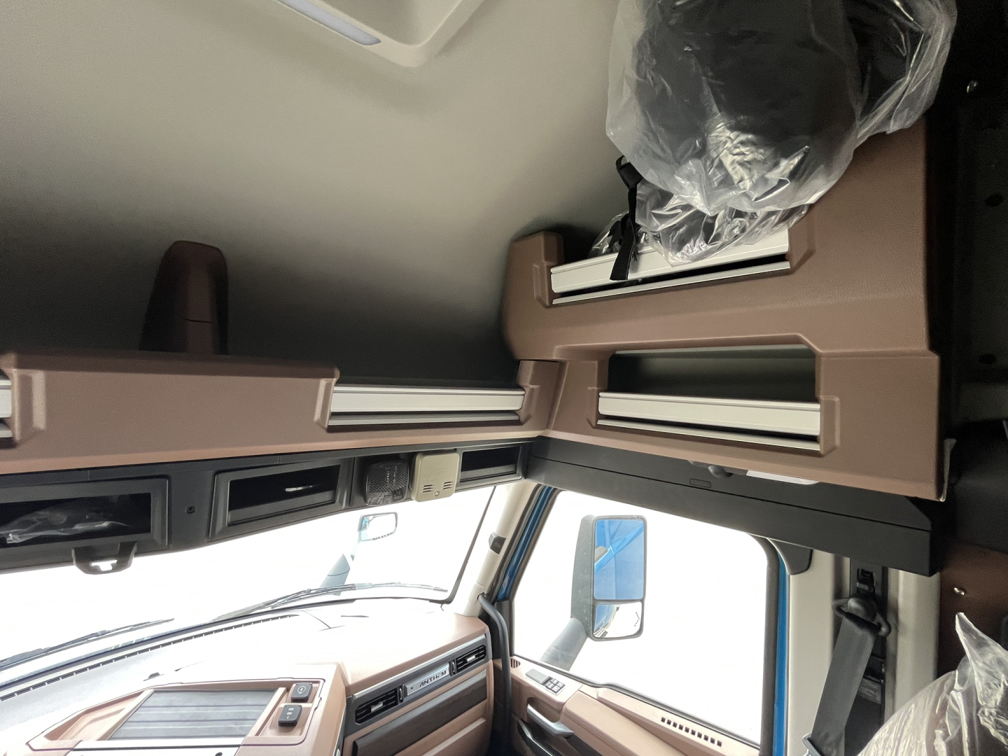 Cab interior of a new Mack AN64T64 truck showing tan dashboard and overhead storage shelves with protective plastic on components.