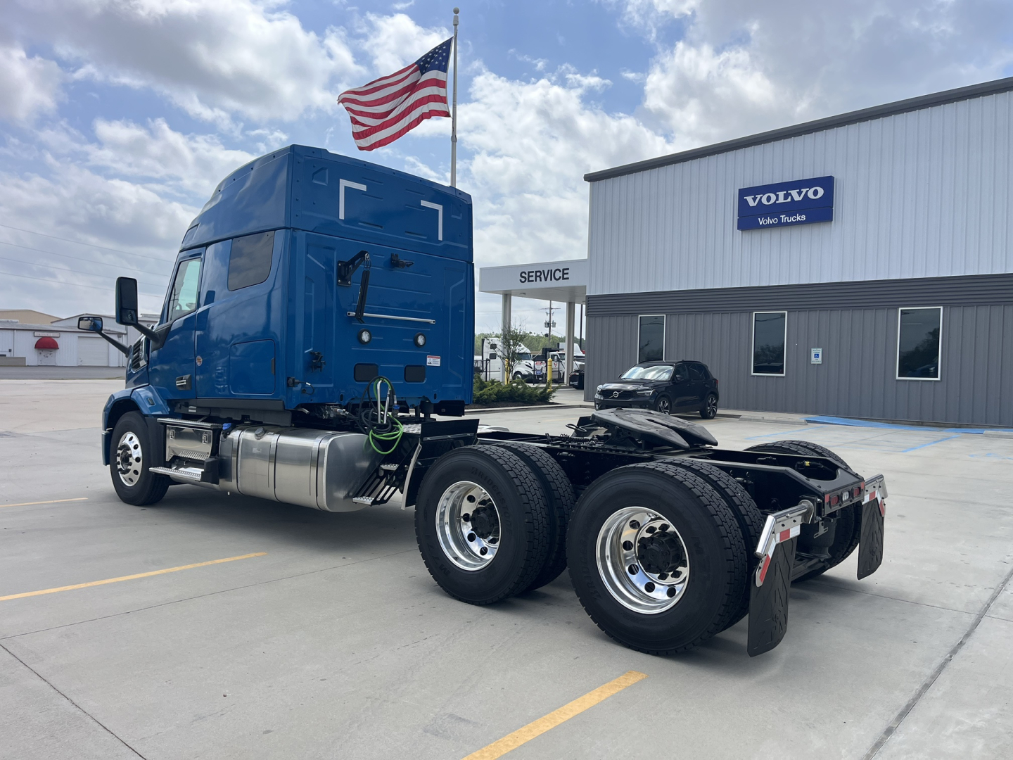 Truck chassis with blue sleeper cab, dual rear axles, parked outside Volvo Trucks dealership; USA flag in background, 2027 Mack AN64T64 listing.
