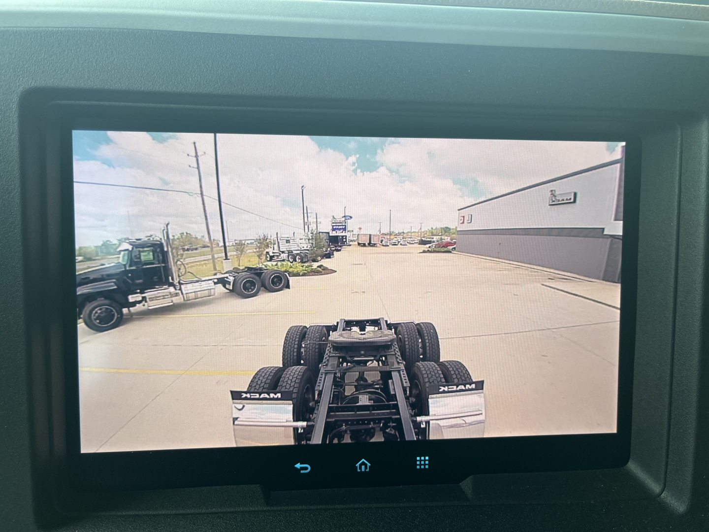 Truck chassis and view from cab of a new Mack AN64T64 conventional sleeper, showing rear drive axles, fifth wheel, and dealership lot in background.