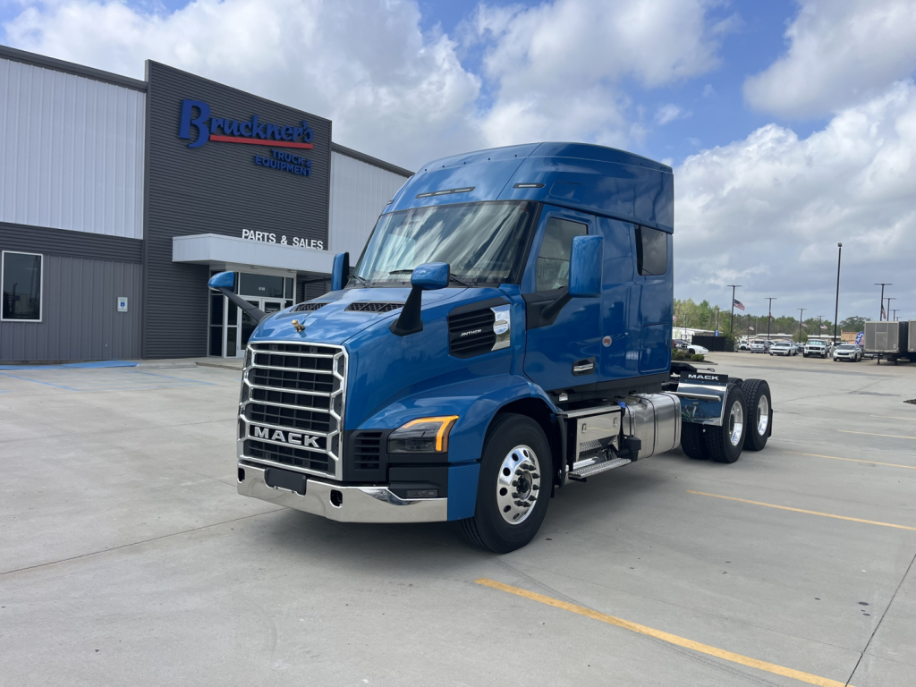 Blue 2027 Mack AN64T64 conventional sleeper truck in a dealership lot, front-right angled view showing chrome grille, Mack emblem, side mirrors, and dual rear axles.