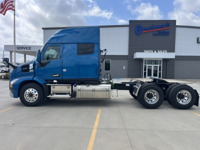 Truck chassis and cab in blue, side view, 6x4 tractor with sleeper cabin, fuel tanks and exhaust visible, parking lot in front of a dealership.