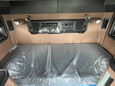 Cab interior of a new Mack AN64T64 truck showing the sleeper cab area with brown dashboard, control panel, and plastic-covered bunk. Visible vents and switches on both sides of the dash.