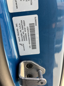Mack Trucks label on blue truck door showing VIN and stock details: 'MACK TRUCKS INC.', 'MACK TRUCKS', 'AVAIL', barcode, and VIN 1M1A24GY8VM102521 with stock number 72145.