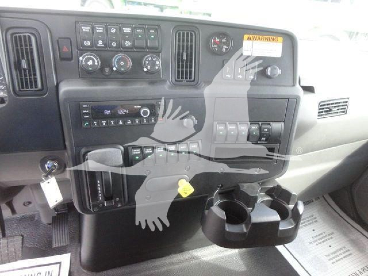 Dashboard and center console of a 2027 International MV truck showing control panels, climate controls, radio, cup holders, and warning sticker.