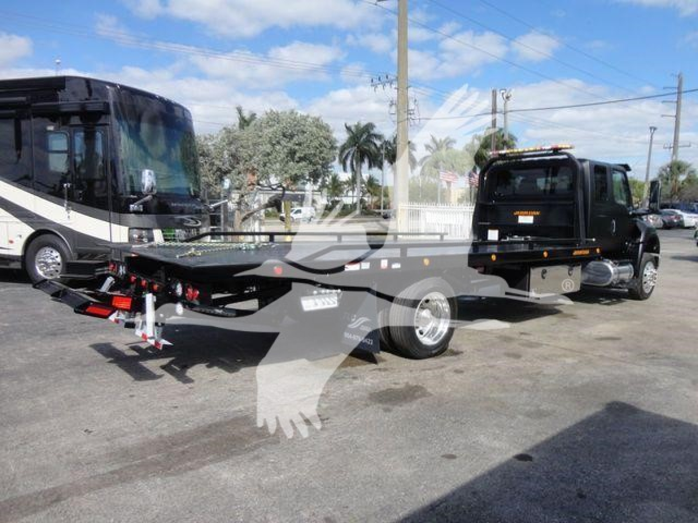 Flatbed tow truck with rollback trailer attached, International MV in background, black tow truck cab, rear hydraulic equipment visible on trailer, outdoor lot.