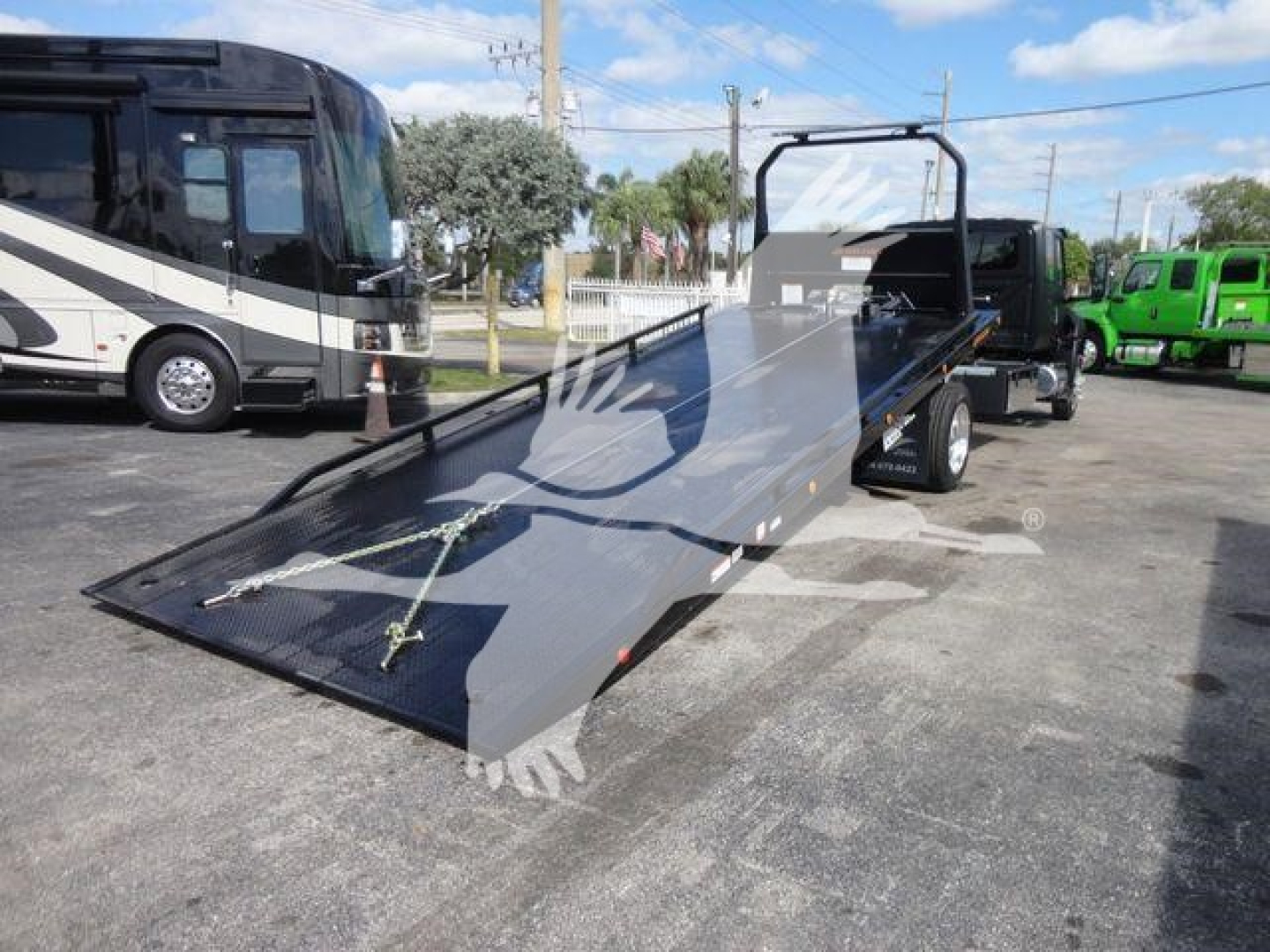 International MV truck with a flatbed rollback trailer in a dealership lot, black tow vehicle, and a storage bus in background.