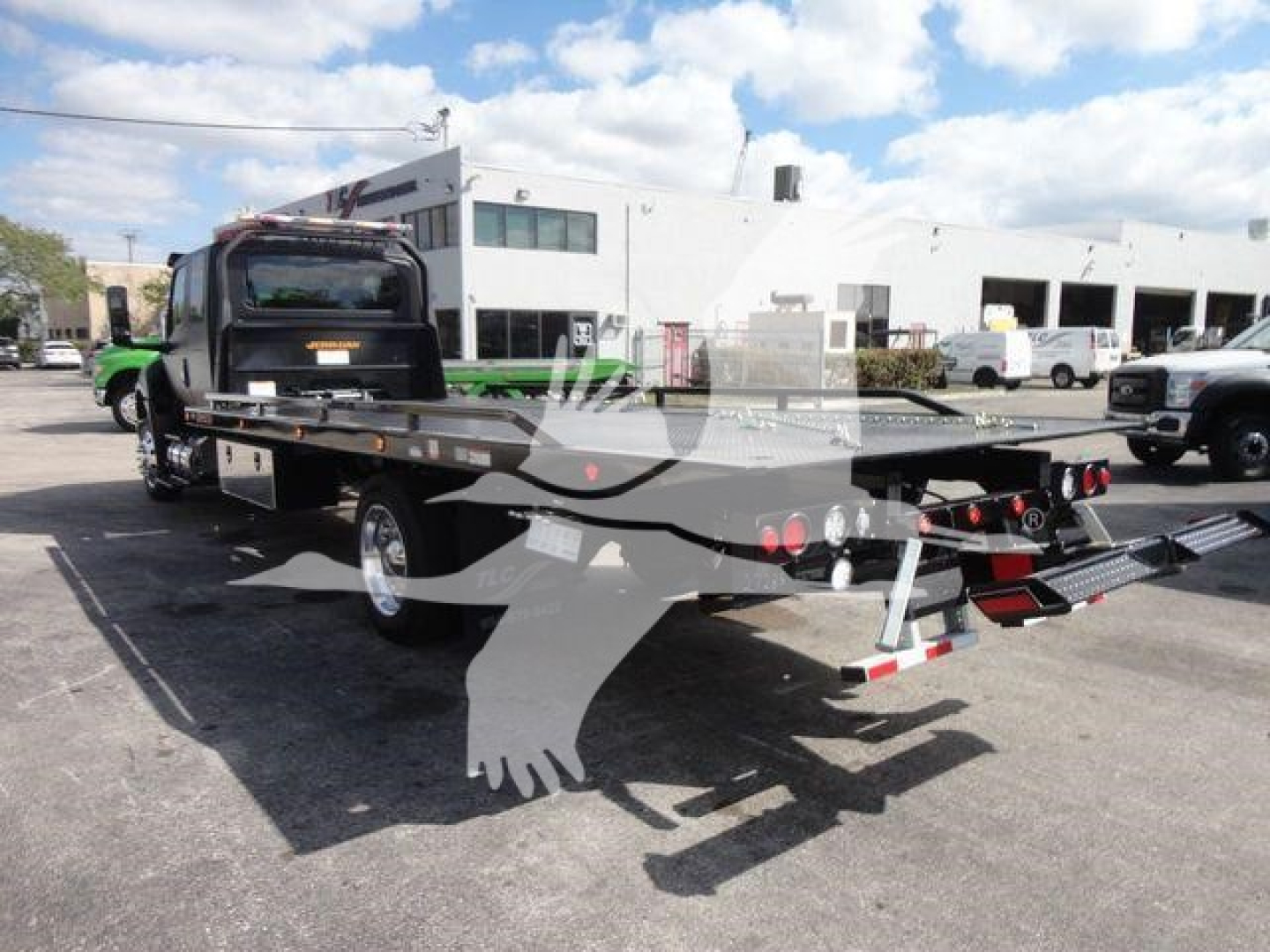 Flatbed rollback tow truck with hydraulic bed in an industrial lot; visible text on truck components but no legible model numbers.
