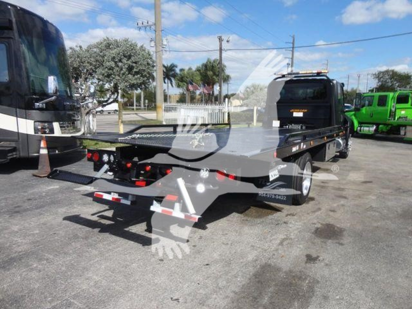 Flatbed tow truck with loading ramps and a black cab, viewed from rear-left side showing equipment bed and lights.