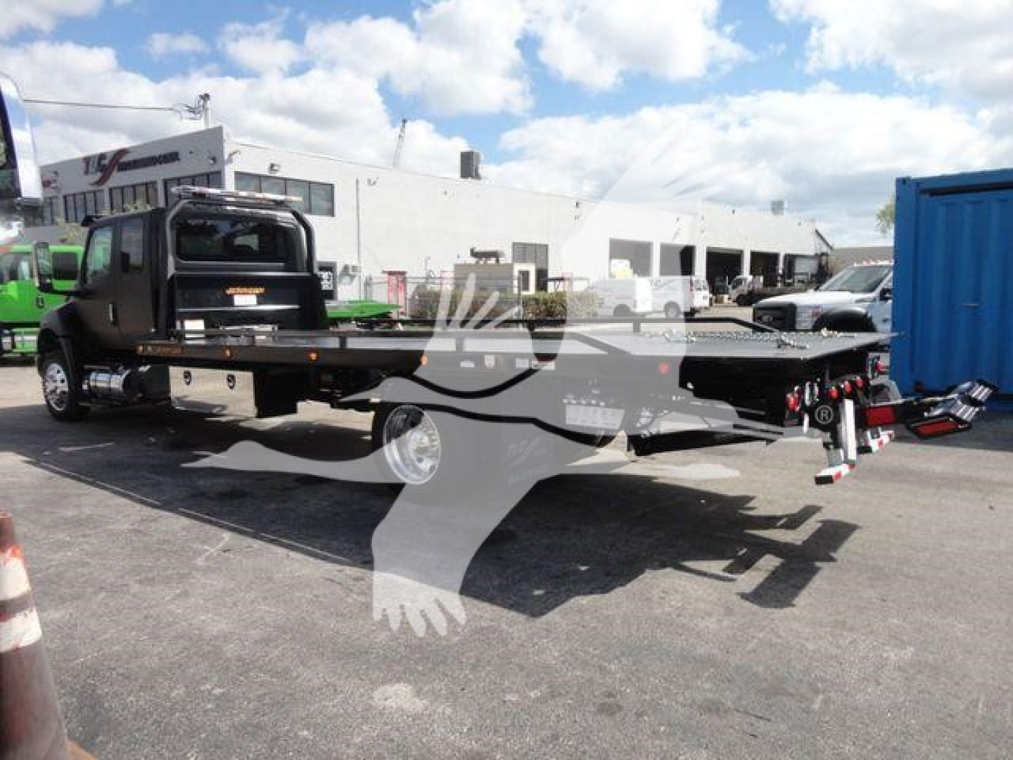 Flatbed tow truck in a dealership lot showing a long flatbed with wheel chocks and lights.