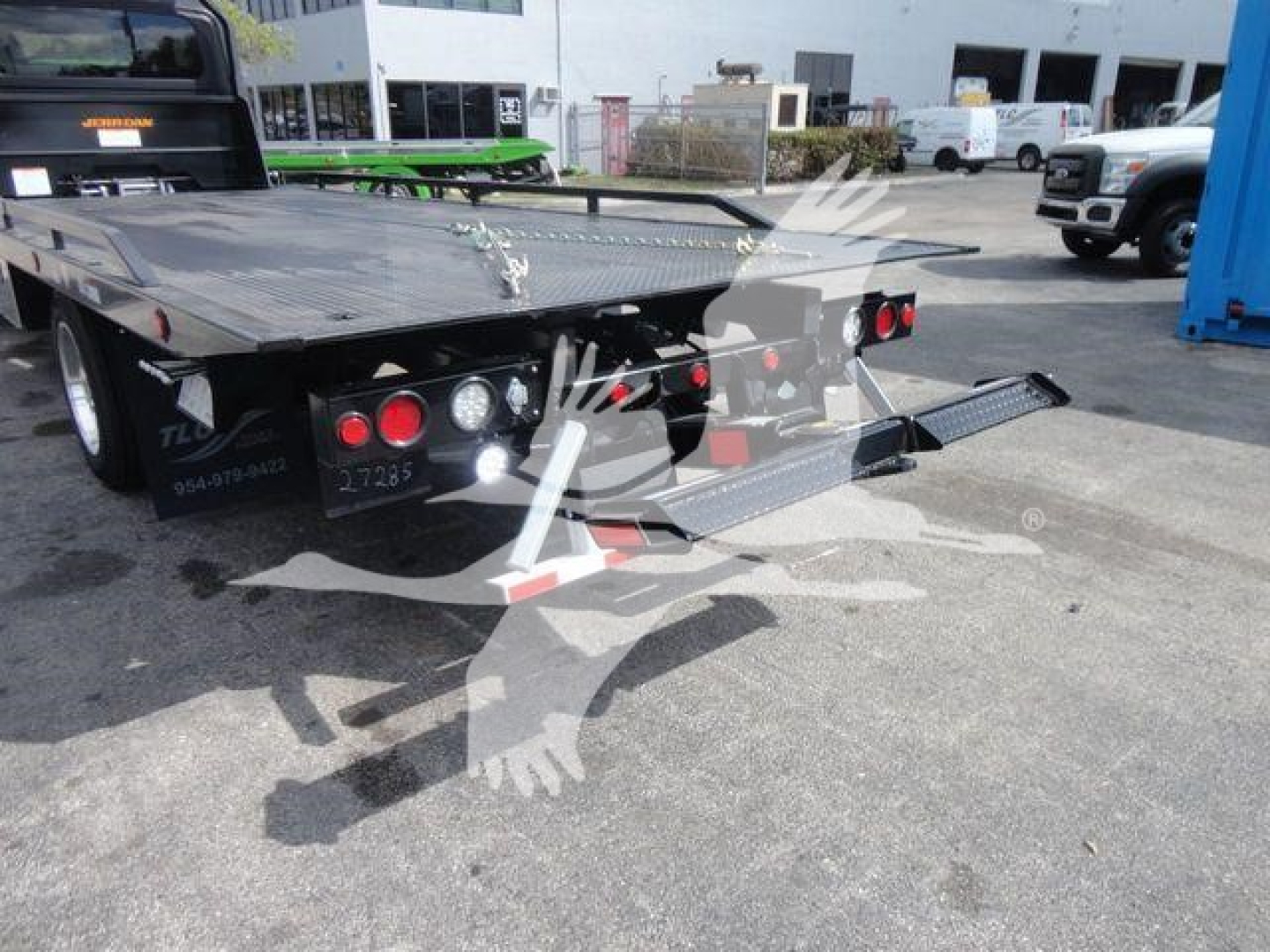 Flatbed tow truck or rollback trailer showing rear ramps and hydraulic controls, with taillights and jack stands visible.