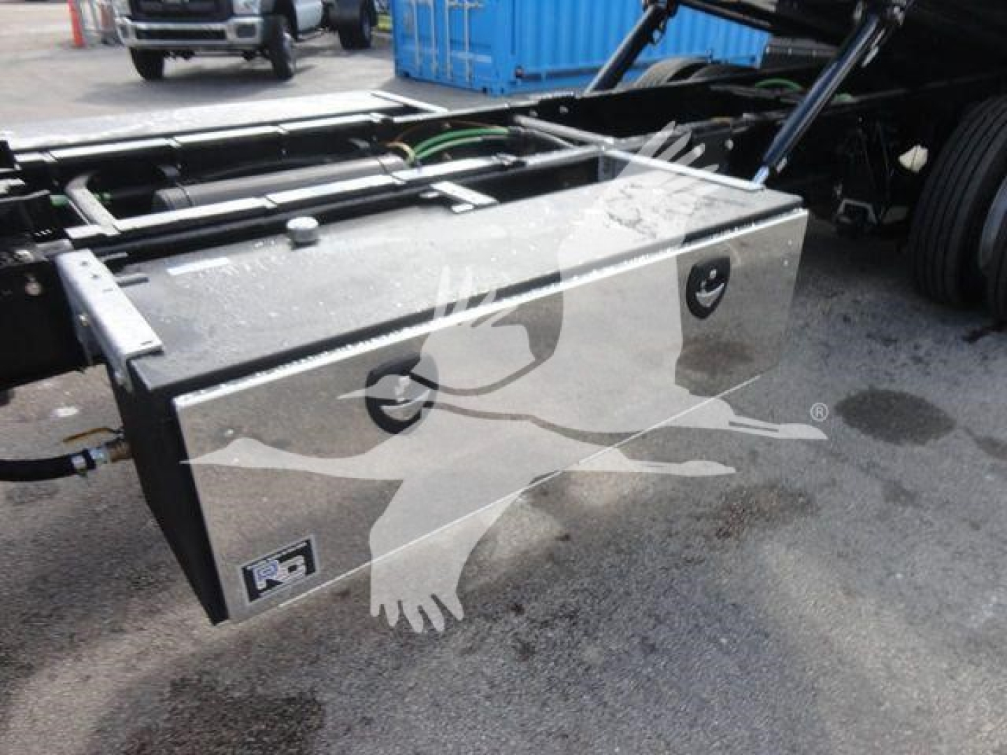 Truck chassis focusing on a rectangular metal toolbox mounted under the cab with hydraulic lines nearby.