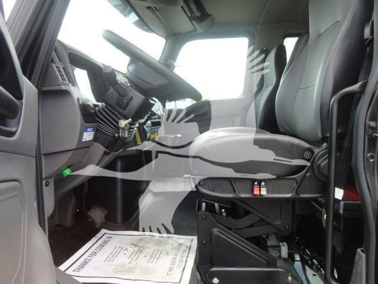 Truck cab interior with driver seat and steering wheel visible; dashboard and pedals; printed paper or mat on floor mat.