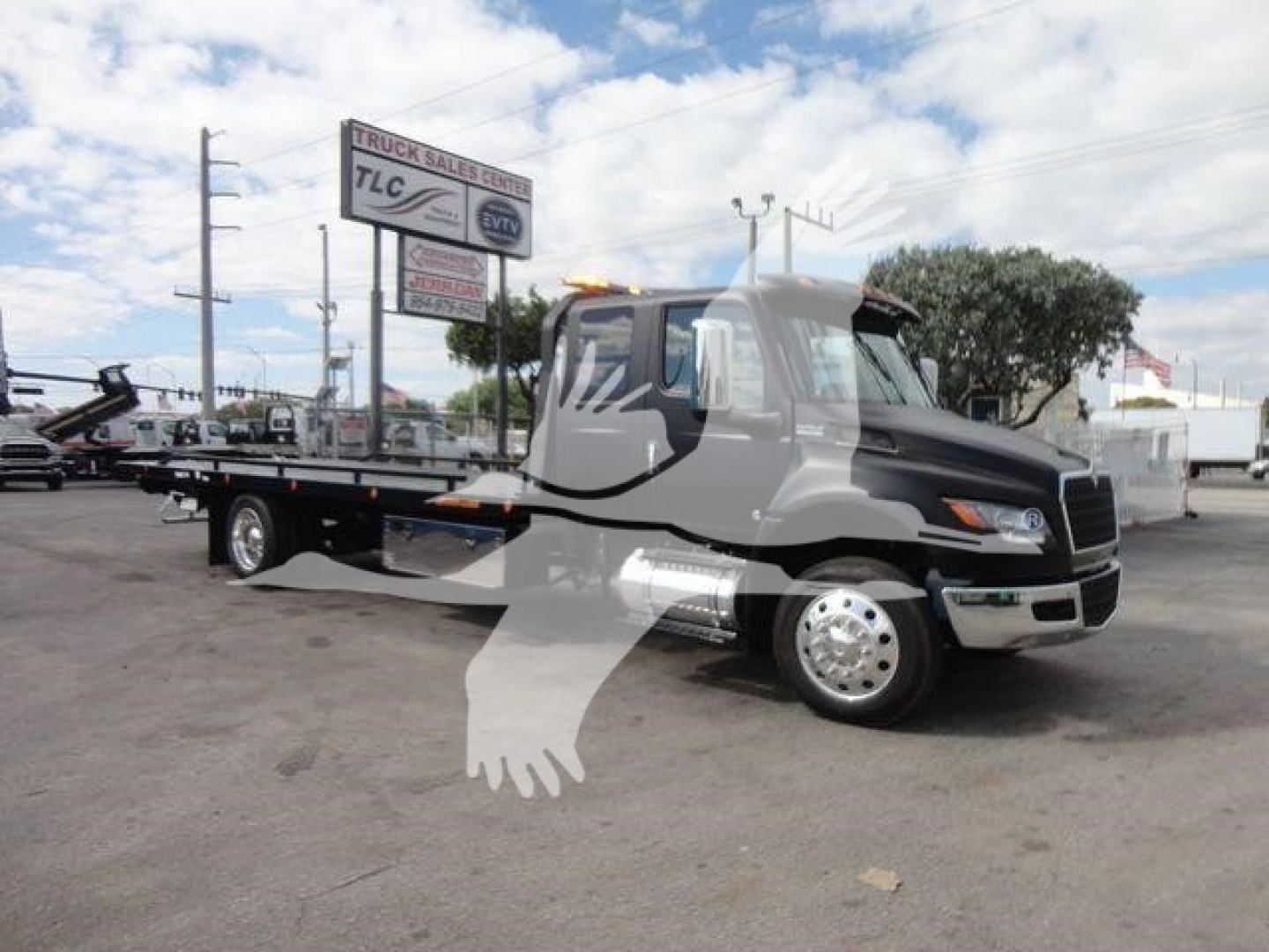 Tow truck with flatbed in a dealership lot showing International MV cab, rollback truck, with sign in background. Visible text on sign includes 'TLC' and 'TRUCK SALES' along with dealer sign.