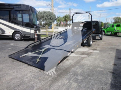 International MV truck with a flatbed rollback trailer in a dealership lot, black tow vehicle, and a storage bus in background.