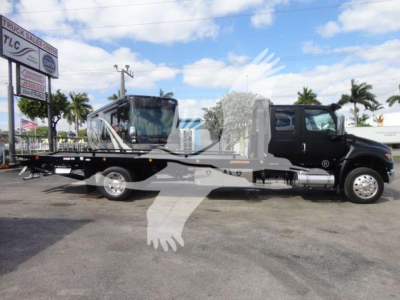 Flatbed tow truck in a dealership lot with palm trees in background and bright sky showing a black cab and long flatbed on a heavy-duty truck.