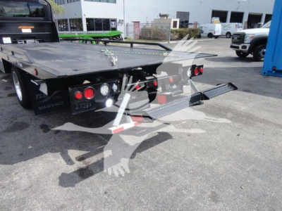 Flatbed tow truck or rollback trailer showing rear ramps and hydraulic controls, with taillights and jack stands visible.
